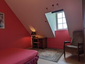 Un dormitorio con techo inclinado, una cama, una silla, un escritorio con lámpara y una ventana.