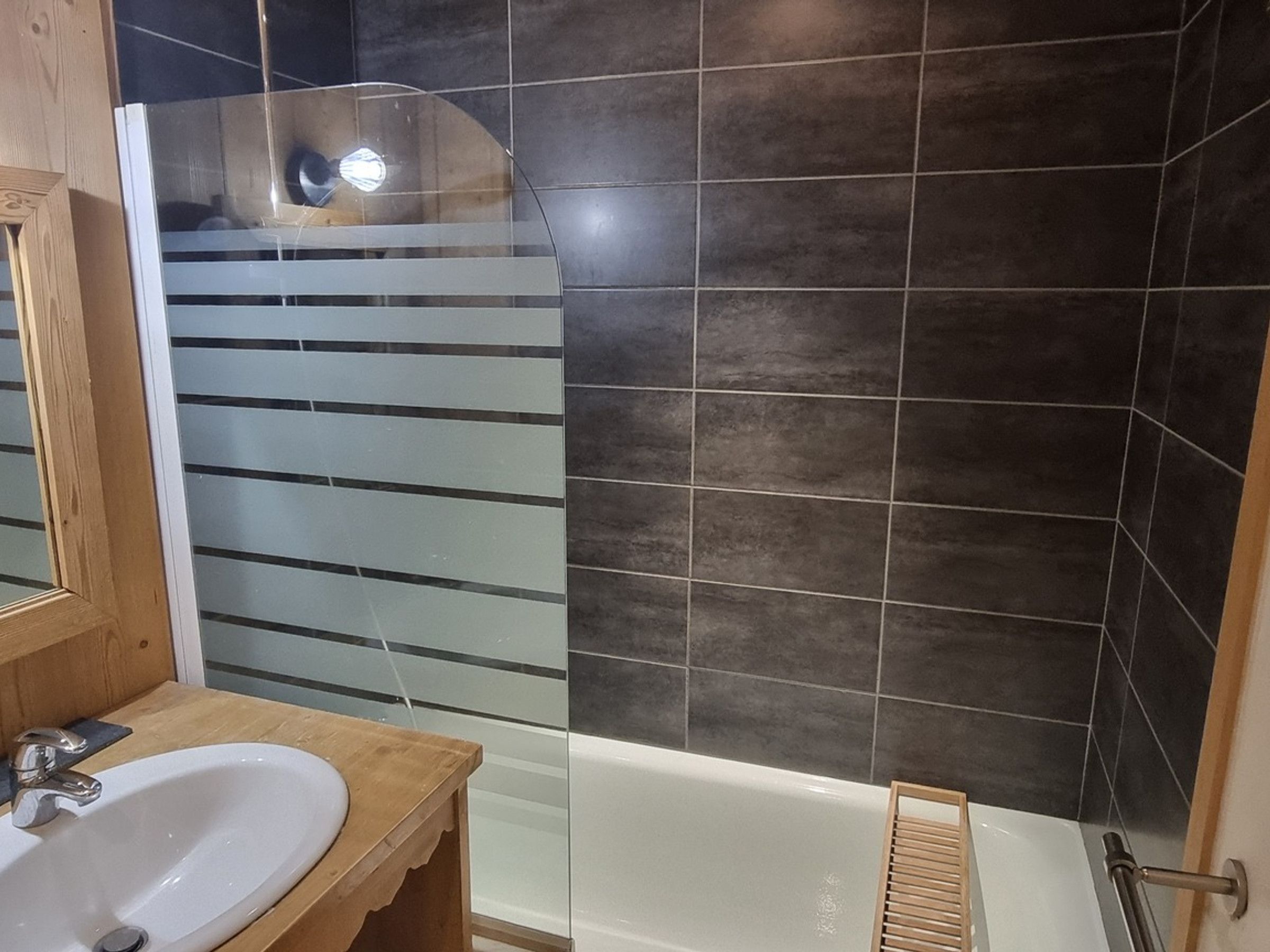 Une salle de bain avec un meuble-lavabo en bois, un lavabo blanc, une cabine de douche en verre et des murs recouverts de carreaux foncés.