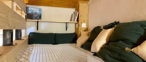 A wooden bunk bed with a metal frame, a mattress with a white grid, and a wall-mounted shelf with books and a framed picture.