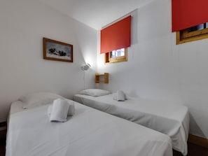 Habitación de hotel con dos camas, una mesita de noche, una lámpara, un ventanal con persianas rojas y un cuadro en la pared.