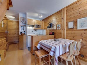 Una habitación acogedora de estilo cabaña, con paredes y suelos de madera, una pequeña cocina americana, un comedor con mantel a rayas y un banco.