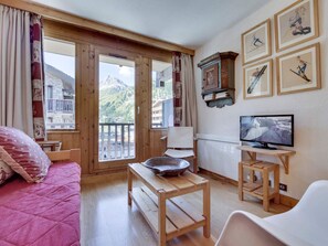 Ein gemütliches Zimmer mit Holzboden, einem Bett mit rosa Bettdecke, einem Holztisch mit Schale, einem weißen Stuhl, einem Fernseher und Blick auf Berge durch große Fenster.