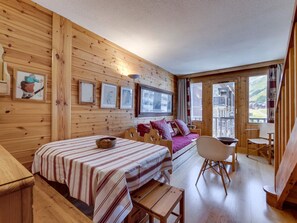 Ein Holzhütten-Innenraum mit einem Bett, einem Tisch und einem Stuhl. An der Wand hängen gerahmte Bilder und ein Fenster bietet einen Ausblick nach draußen.
