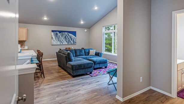 A modern living room with a sectional sofa, a dining area with chairs, a colorful rug, and a large window providing natural light. - Full Duplex Staging Ground for Large Family Groups (Girdwood)