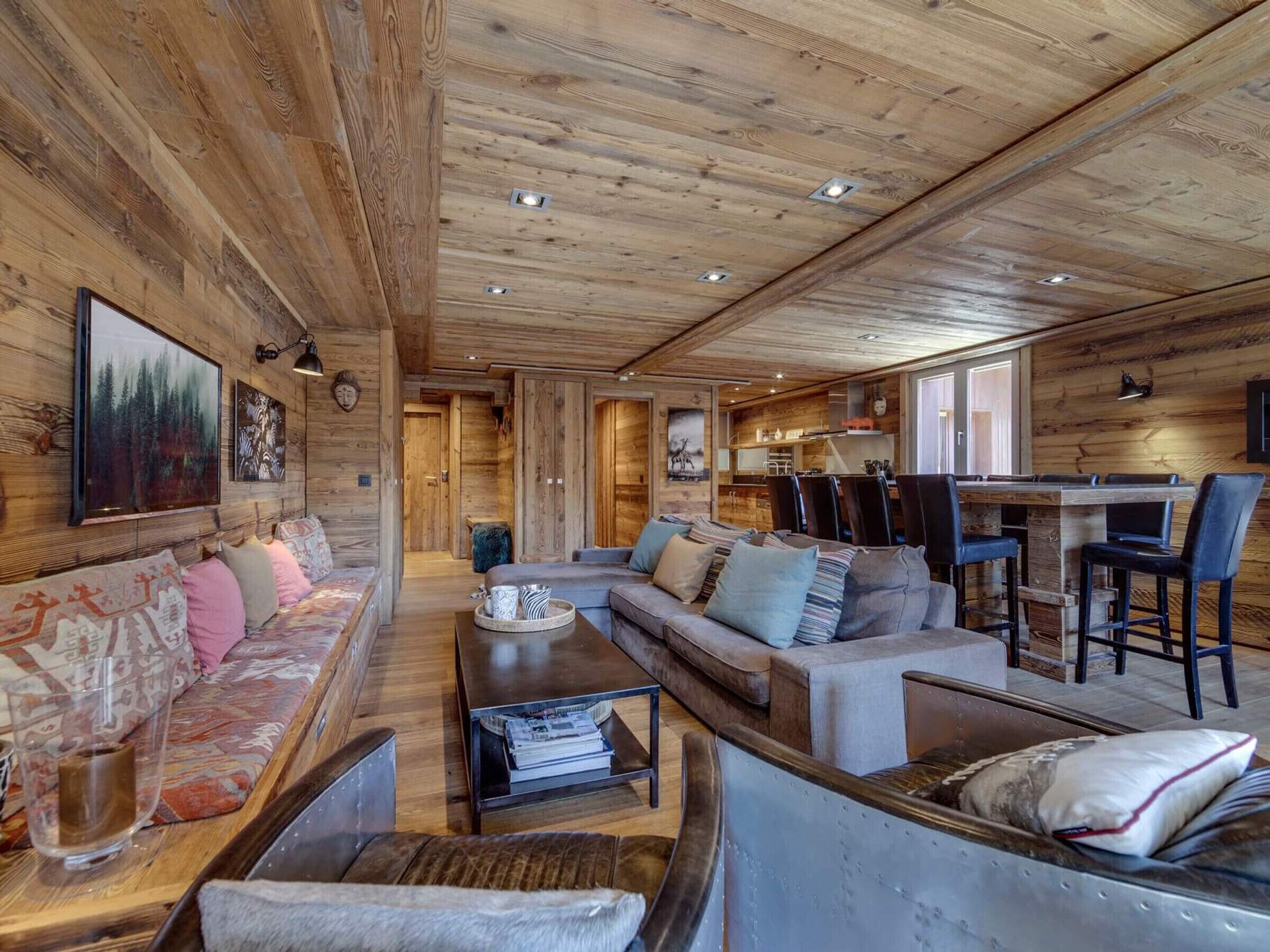 Ein gemütliches Blockhaus-Interieur mit Holzbalkendecke, einer Sofagarnitur, einem Essbereich und einer durch eine offene Tür sichtbaren Küche.