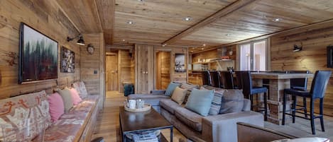 A cozy cabin interior with a wooden ceiling, a sofa set, a dining area, and a kitchen visible through an open door.