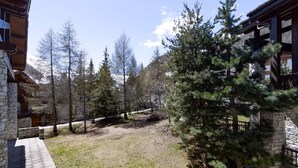 A cabin with stone walls, a wooden deck, and a view of a forested area with tall trees. - Chalet apartment, central quiet area - 8 guests (Val-d'Isère)