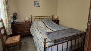 Ein Schlafzimmer mit einem Einzelbett, einem Holznachttisch, einem Sessel, einem Ventilator und einer Wandleuchte.
