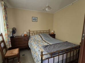 Ein Schlafzimmer mit einem Einzelbett, einem Holznachttisch, einem Sessel, einem Ventilator und einer Wandleuchte.