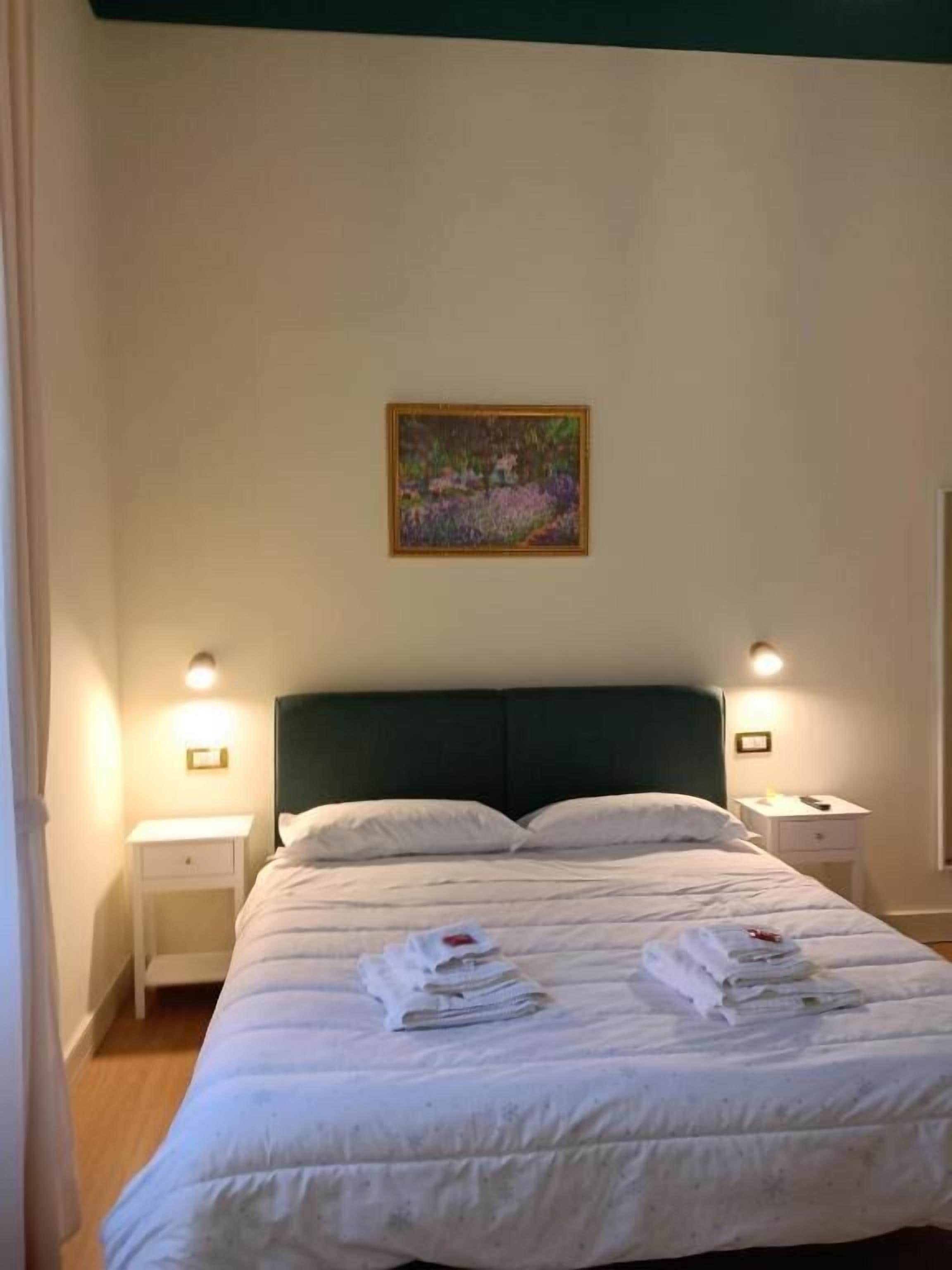 Een netjes opgemaakt bed met witte lakens en handdoeken, twee nachtkastjes met lampen, en een schilderij aan de muur.