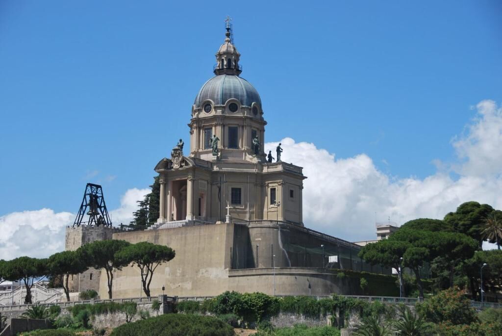 Um edifício histórico com cúpula e estátuas, rodeado por árvores e um céu claro.