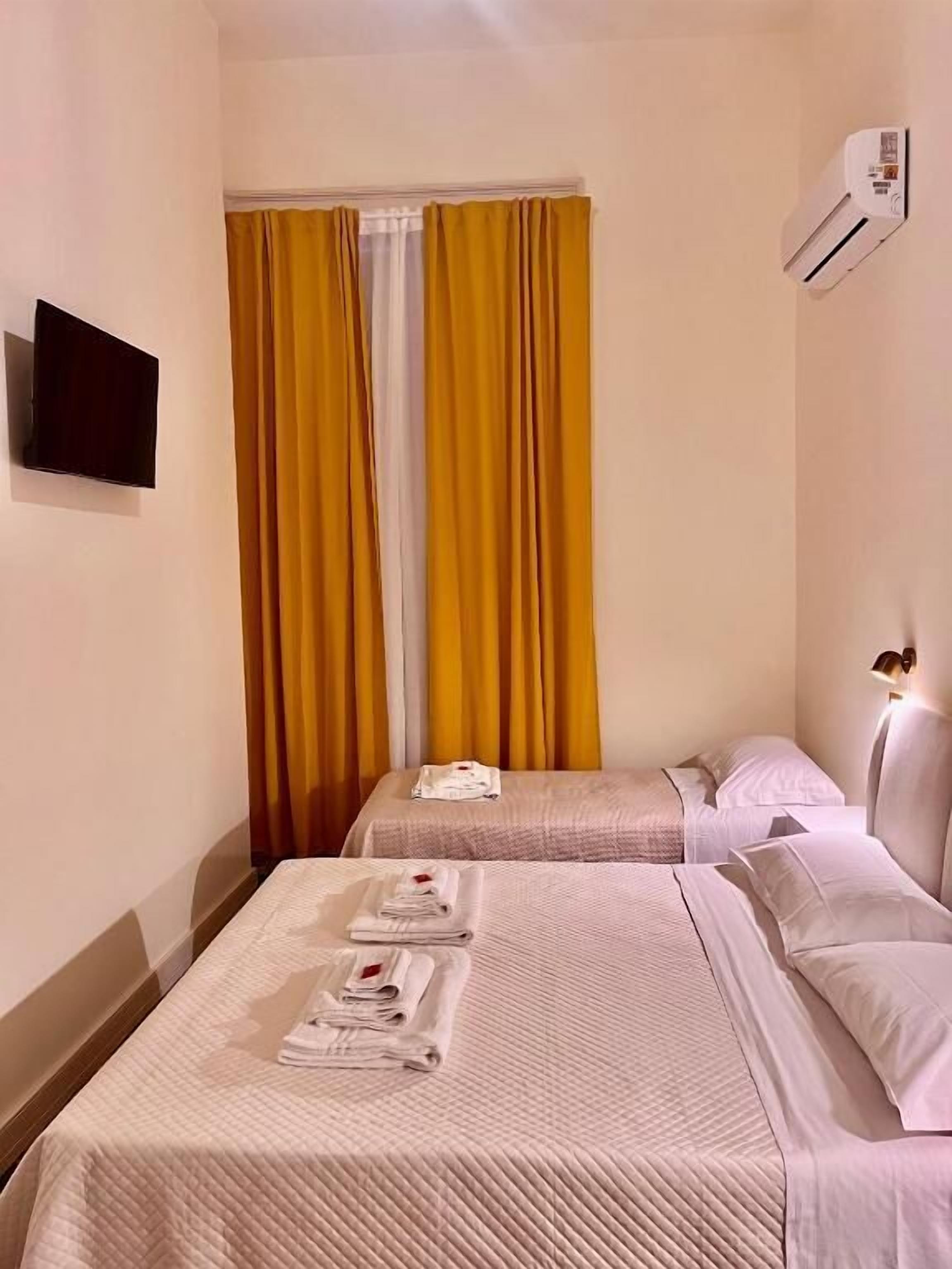 Une chambre d’hôtel avec deux lits, un téléviseur à écran plat, un appareil de climatisation et une fenêtre avec des rideaux jaunes.