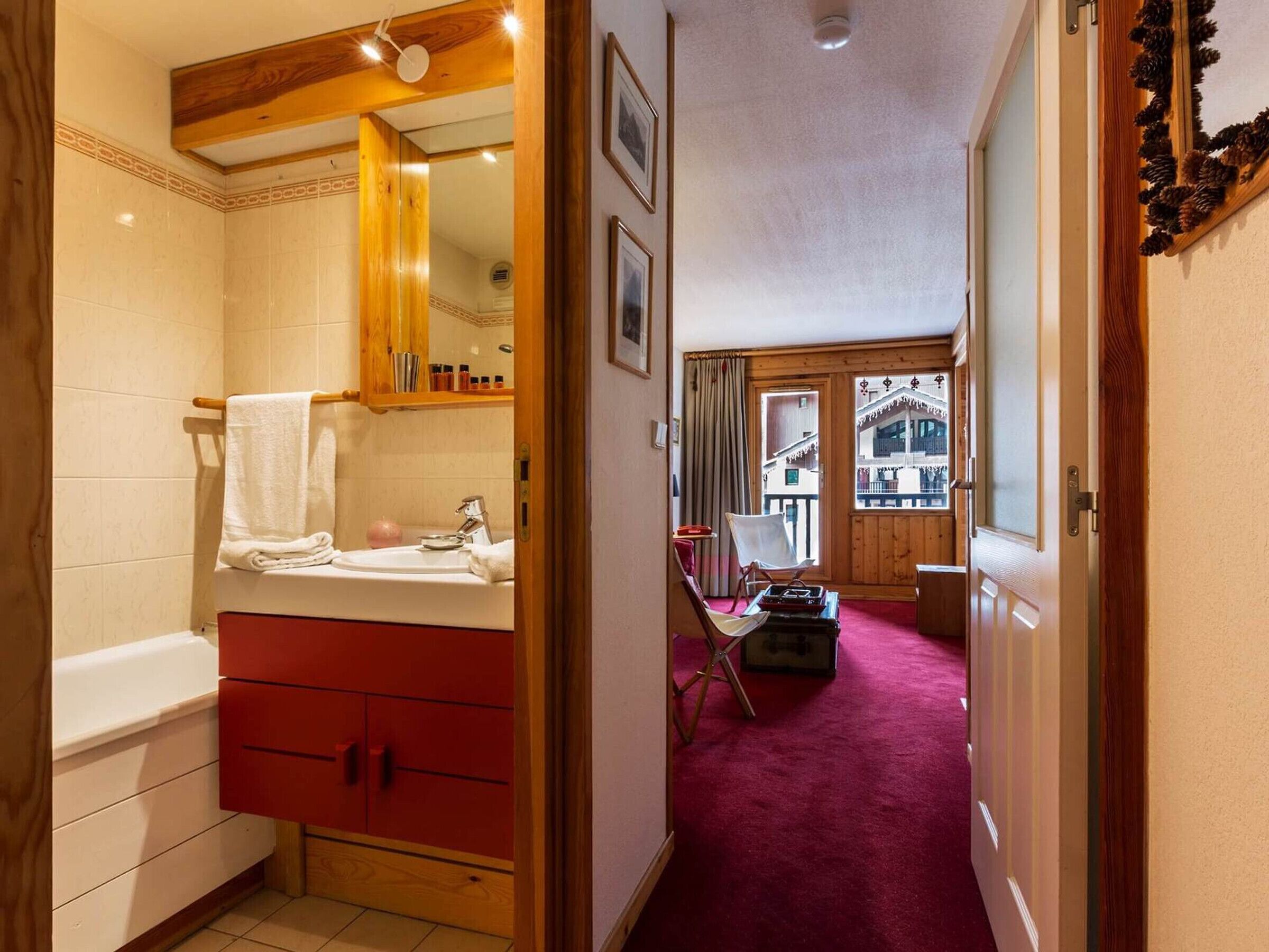 Un baño con un armario rojo, un lavabo blanco y un espejo. Un tapete rojo conduce a un balcón con una silla y una maleta.