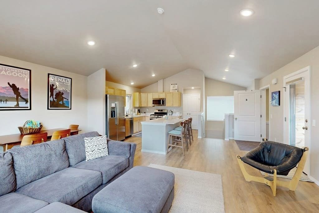 A modern living room with a grey sectional sofa, a black leather chair, and a kitchen area with wooden cabinets and a central island.