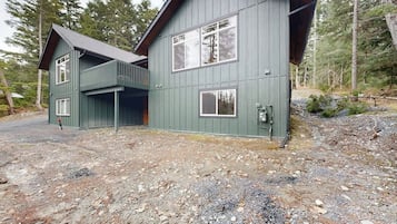 A two-story cabin with a balcony, surrounded by a forested area.