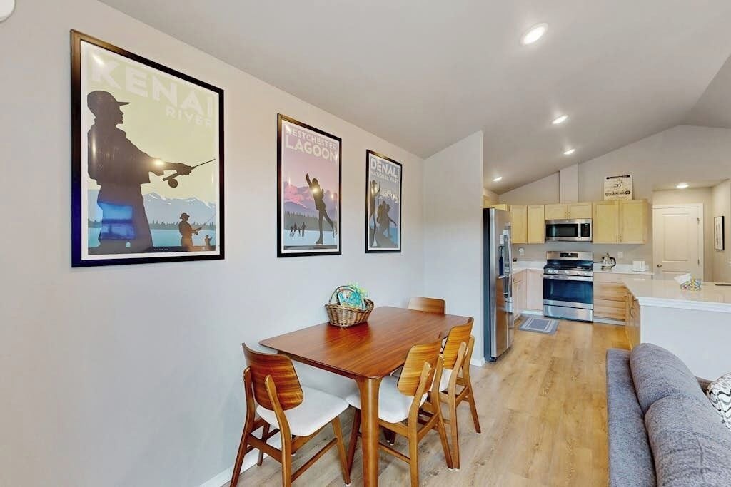 A dining area with a wooden table and chairs, a kitchen with modern appliances, and framed pictures on the wall.