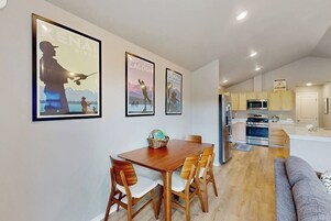 Un comedor con una mesa y sillas de madera, una cocina con electrodomésticos modernos y cuadros en la pared.