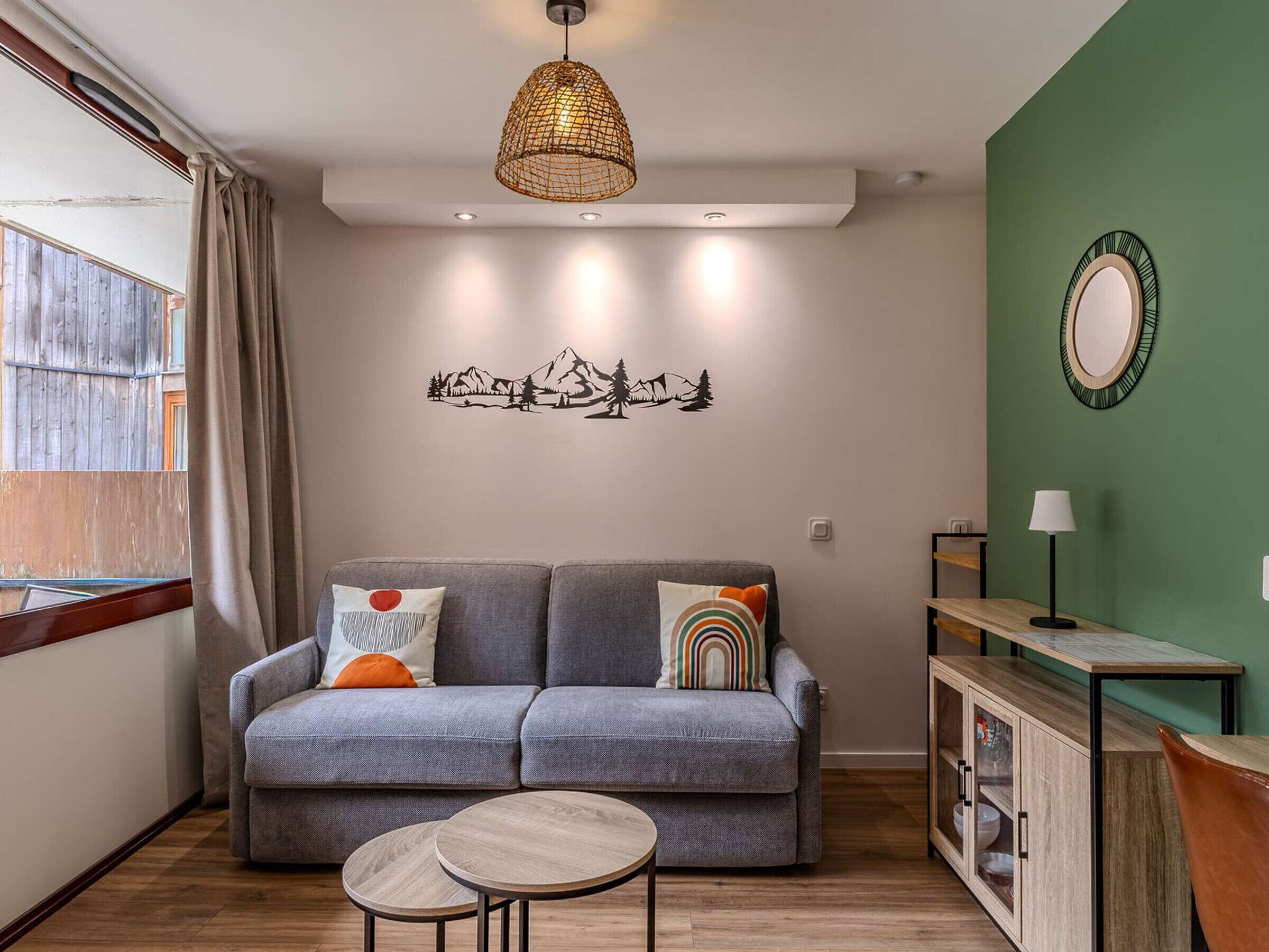 A modern living room with a grey sofa, two round wooden side tables, a woven pendant light, a wall-mounted mirror, and a green accent wall.
