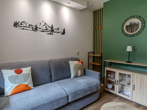 A living room with a grey sofa, a wall decal of mountains, a green accent wall, a wooden cabinet with glass doors, and a round mirror. - Cozy apartment near slopes - 4 guests (Plagne Bellecôte)