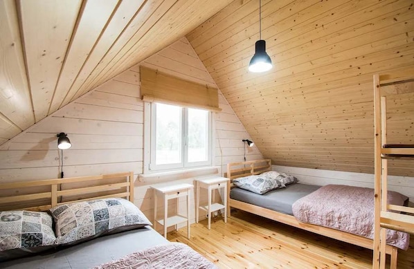 A dormitory room with wooden bunk beds, a desk, and a window.