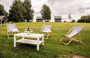 A grassy outdoor area with a white table set with wine glasses and a bottle, two lounge chairs, and a small wooden structure in the background.