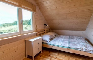 A small, single-bed room with a wooden ceiling, a bedside table, a lamp, and a window with a view of greenery.
