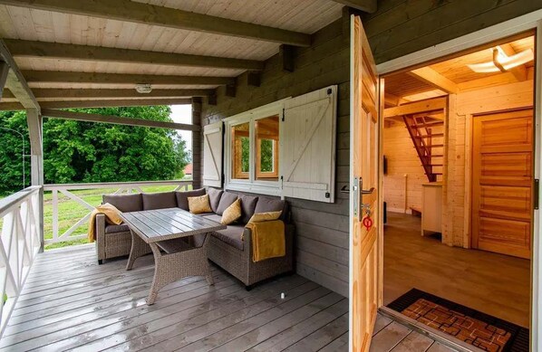 A covered outdoor seating area with a sofa and table, wooden walls, and a staircase leading to an interior room.