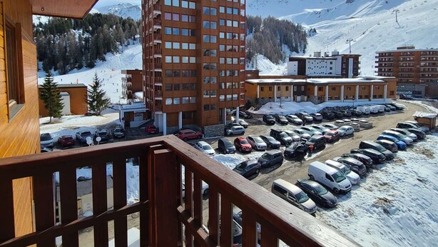 Um edifício alto com vários andares, um estacionamento com diversos carros e uma paisagem montanhosa nevada ao fundo.