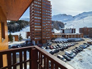 Un immeuble de grande hauteur, avec de nombreux étages, un parking où sont garées plusieurs voitures, et un paysage montagneux enneigé en arrière-plan.