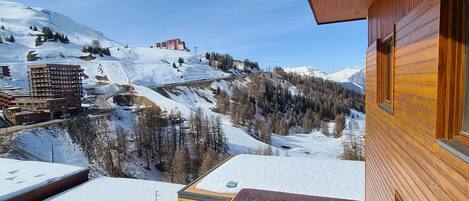 A wooden cabin with a balcony overlooking a snowy mountain landscape, including a ski lift and buildings.