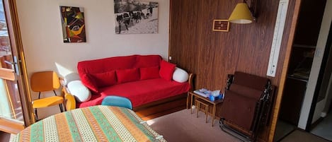 Une pièce avec un canapé rouge, un meuble en bois, une petite table avec une lampe et deux chaises.