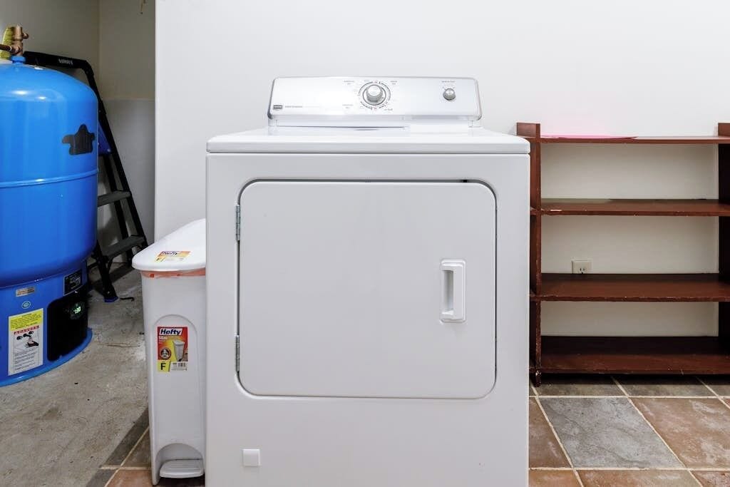A compact, white front-loading washer and dryer unit with a control panel on top, a small trash bin with a Hefty label, and a blue propane tank to the left.