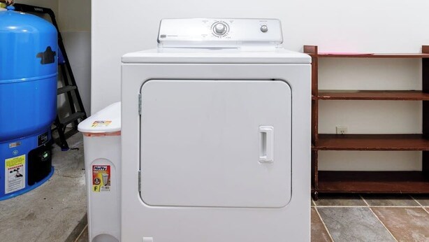 Eine kompakte, weiße Waschmaschine und ein Trockner in einem, mit Bedienfeld oben, eine kleine Mülltonne mit Hefty-Aufdruck und eine blaue Propangasflasche auf der linken Seite.