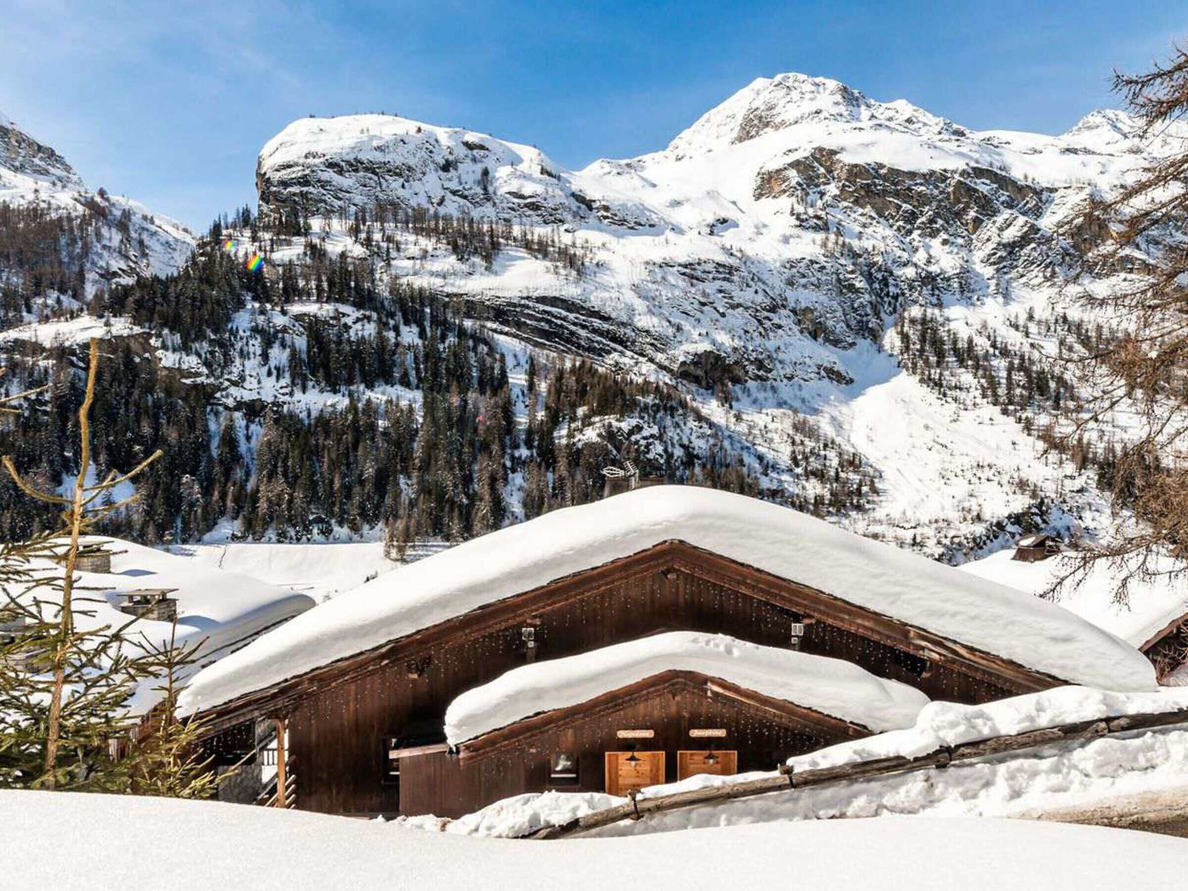 A wooden chalet with a snow-covered roof, surrounded by snow-covered mountains and trees.