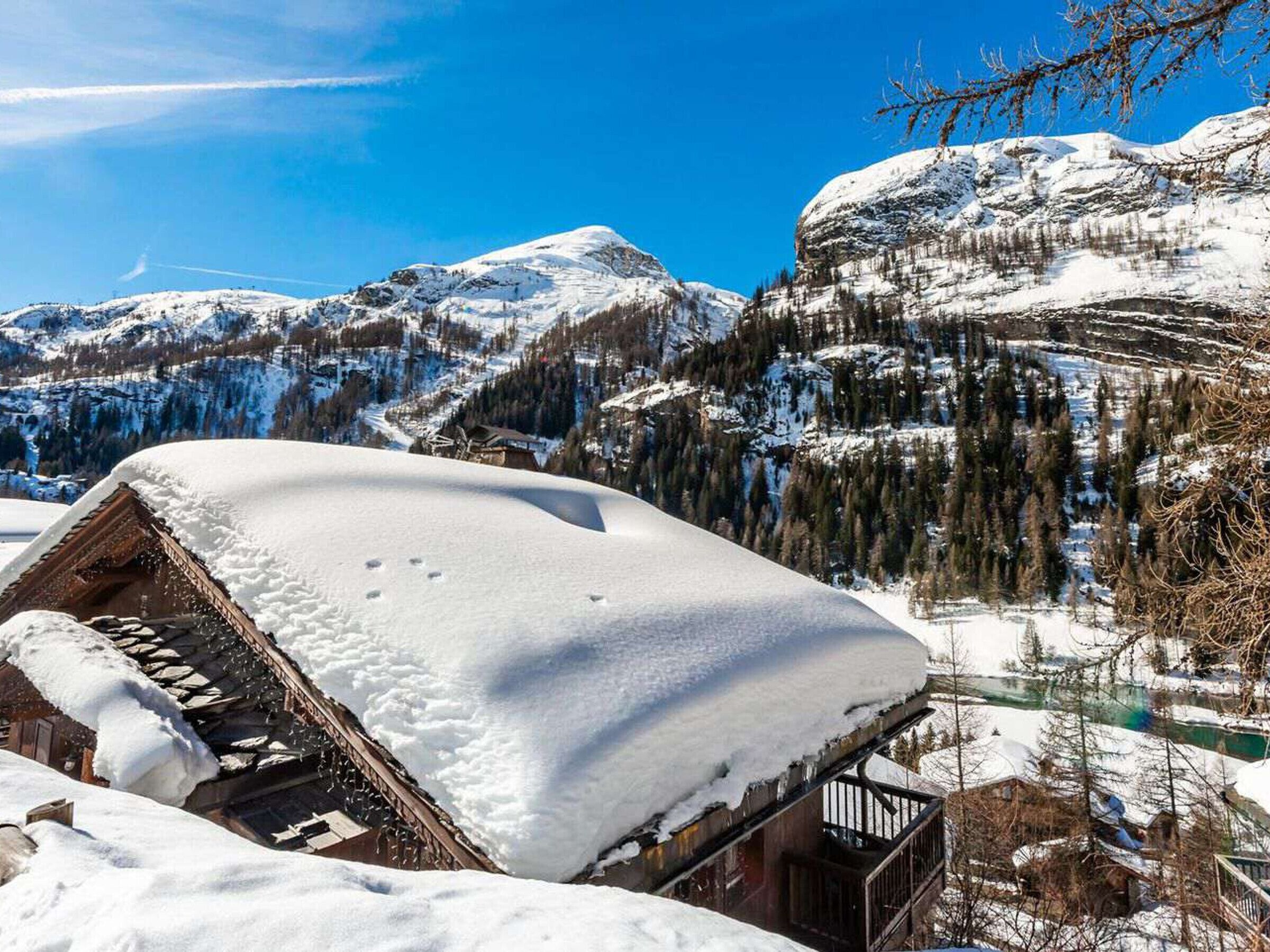 A wooden cabin with a snow-covered roof, surrounded by snow-covered mountains and a clear blue sky.