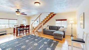 A spacious living area with a wooden staircase, a gray sofa, a dining table with chairs, and a patterned rug.