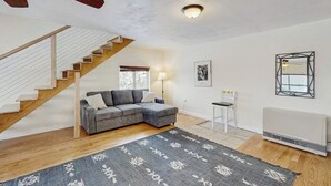 A living room with a staircase, a gray sectional sofa, a patterned rug, a white radiator, and a framed picture on the wall.