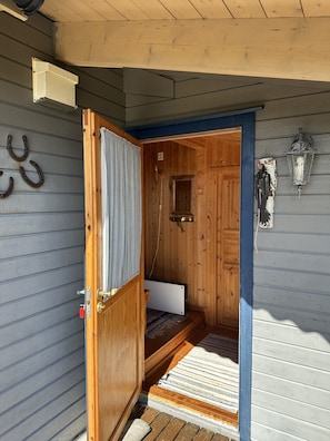 Una cabaña de madera con una puerta que da a una habitación pequeña con un banco, una lámpara de pared y una ventana.