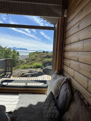 A wooden cabin with a large window overlooking a rocky landscape and a distant mountain range.