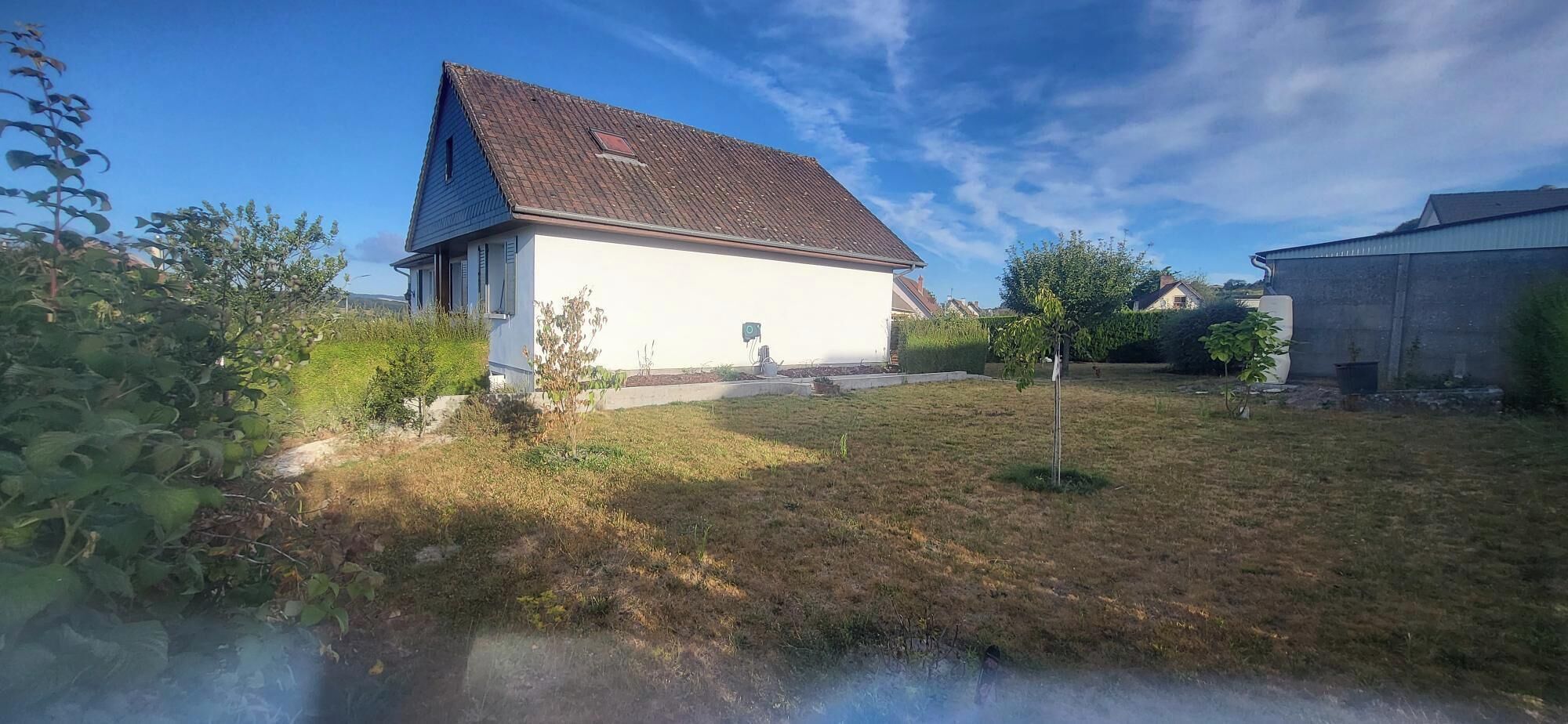 Une maison de plain-pied avec un toit en pente, entourée d’une cour verdoyante et de quelques plantes.