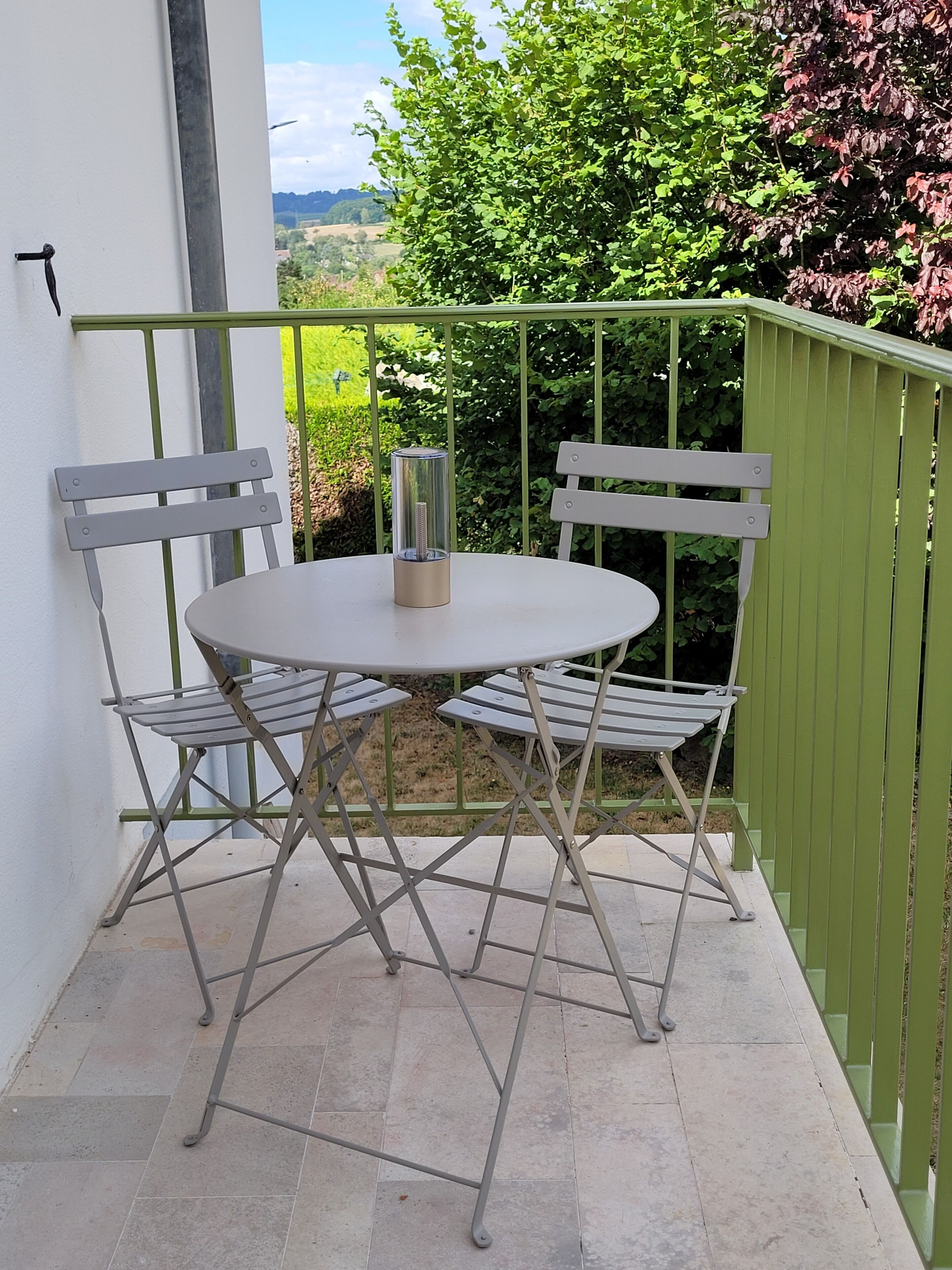 A small outdoor patio with a round table and two chairs, a metal railing, and a view of greenery and hills in the background.