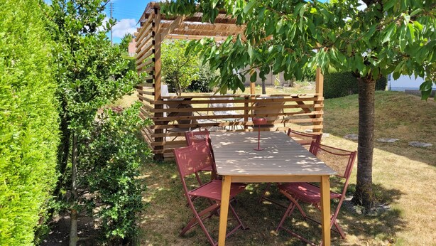 A wooden deck with a table and chairs, surrounded by greenery and a building in the background. - Gîte des Tulipes de Bray (Neufchâtel-en-Bray)