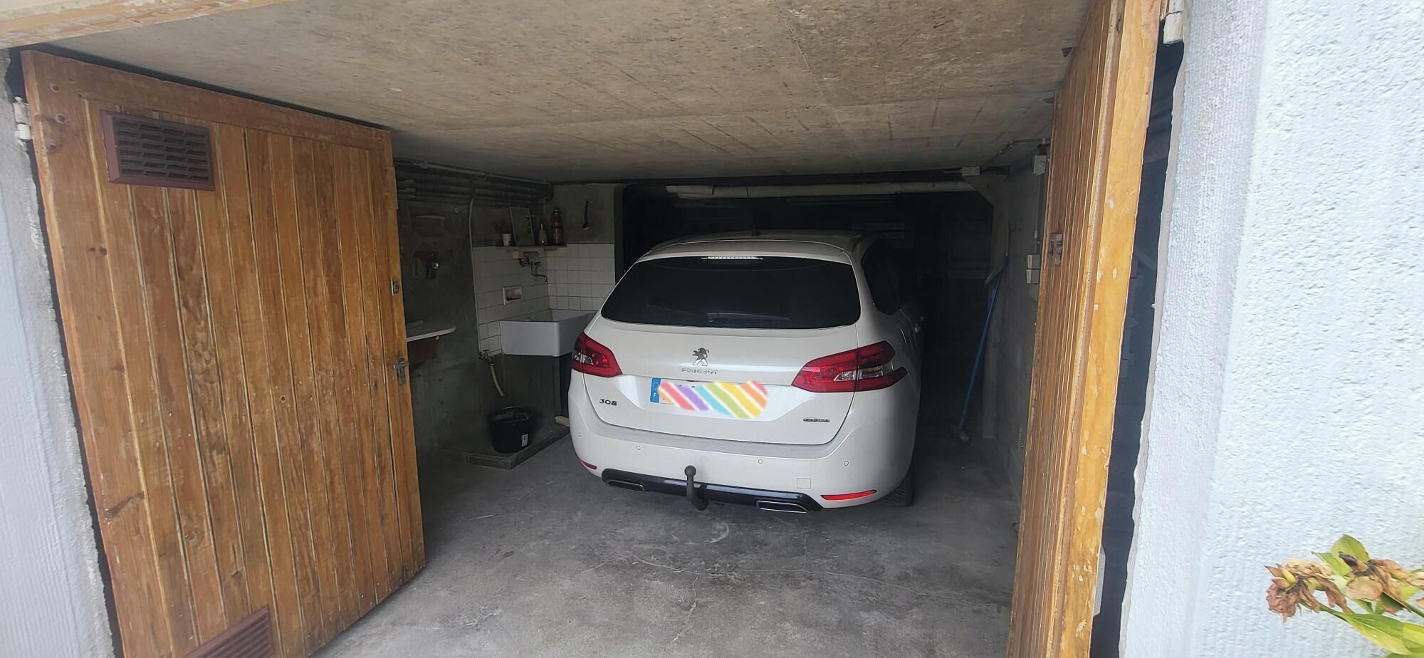 Une voiture blanche est garée dans un garage étroit, doté d’une porte en bois et d’un mur recouvert de carreaux.