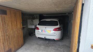 Une voiture blanche est garée dans un garage étroit, doté d’une porte en bois et d’un mur recouvert de carreaux.