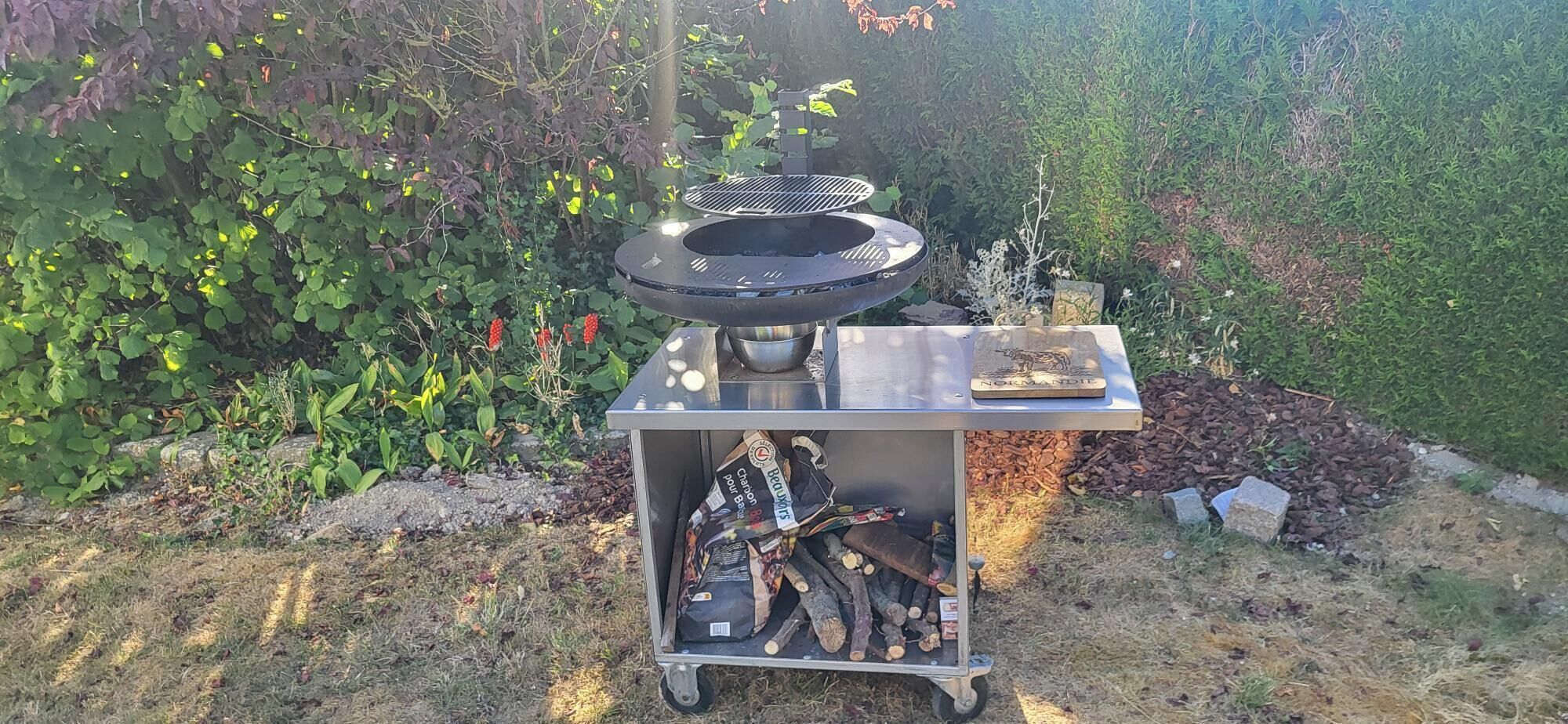 Un barbecue portable doté d’une grille en métal et d’un logo, placé à l’extérieur, avec un jardin en arrière-plan.