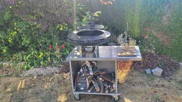 Un barbecue portable doté d’une grille en métal et d’un logo, placé à l’extérieur, avec un jardin en arrière-plan.