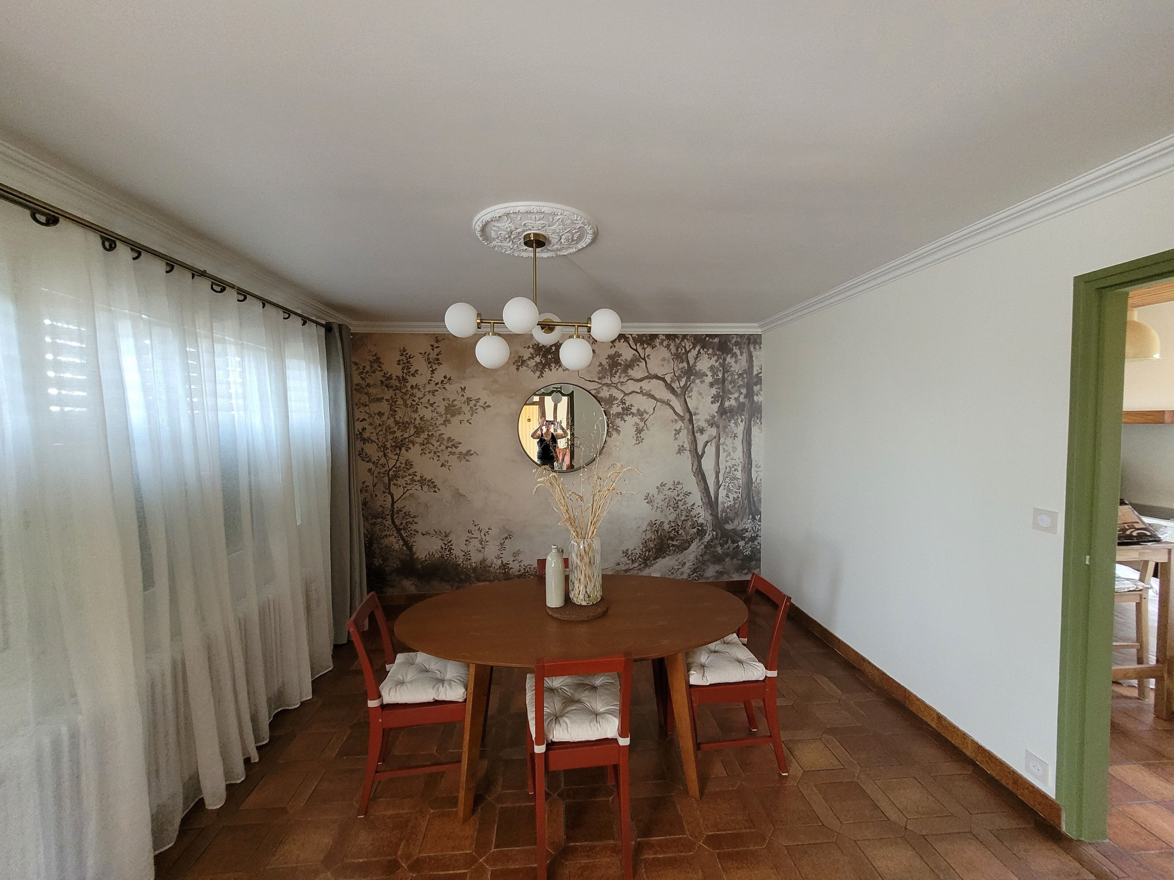 A dining area with a round table, four chairs, a vase with dried plants, and a wall mural.