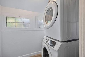 A laundry room with a front-loading washer and dryer, a window with frosted glass, and a white wall. - The Dellwood Cottage - Near Local Shops & Eats (Jacksonville)