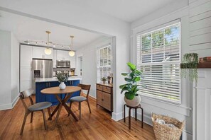 A kitchen with modern appliances, a dining area with a wooden table and chairs, and a large window with blinds. - The Dellwood Cottage - Near Local Shops & Eats (Jacksonville)