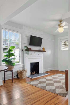 A living room with a fireplace, a television mounted above it, a ceiling fan, and a large window with blinds. - The Dellwood Cottage - Near Local Shops & Eats (Jacksonville)